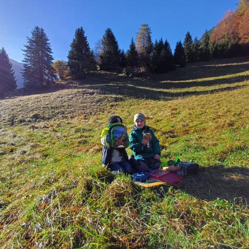 2 Kinder beim Jausnen auf einer Wiese - ein Kind versteckt sich hinter seinem Rucksack