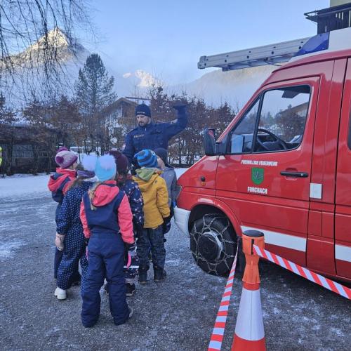 Neben dem Feuerwehrauto stehen eine Feuerwehrmann, ein Polizist und die Volksschulkinder 