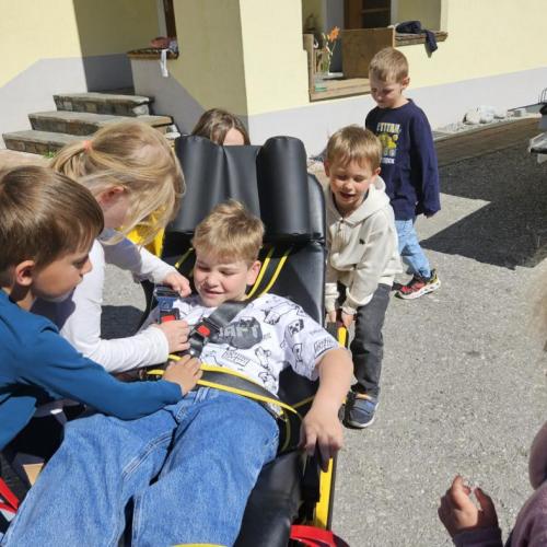 Schüler schnallen einen Mitschüler auf der Liege fest, im Hintergrund ist das Rettungsauto 