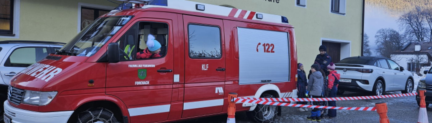 Eine Schülerin sitzt im Feuerwehrauto und mehrere Kinder stehen hinter und neben dem Feuerwehrauto.