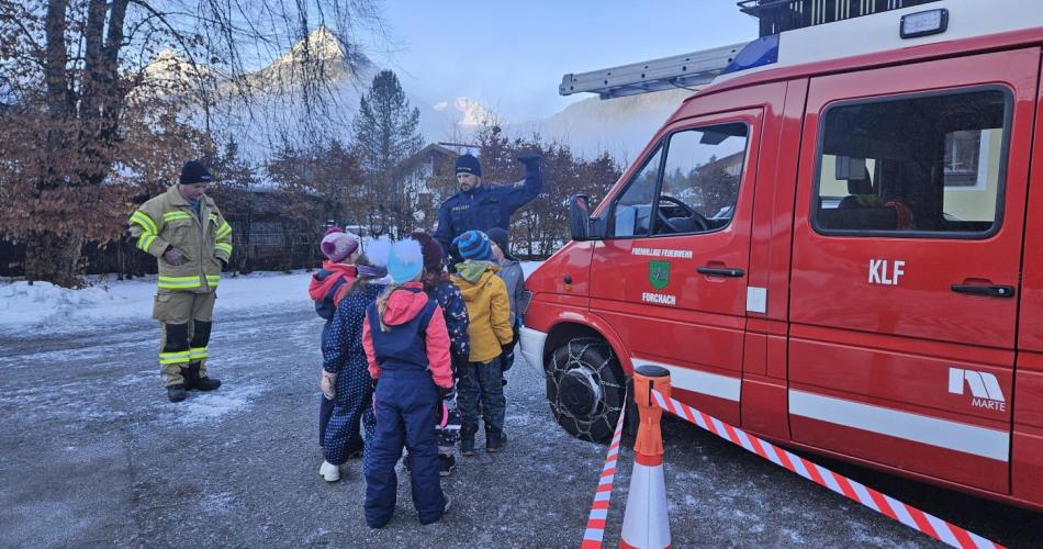 Neben dem Feuerwehrauto stehen eine Feuerwehrmann, ein Polizist und die Volksschulkinder 
